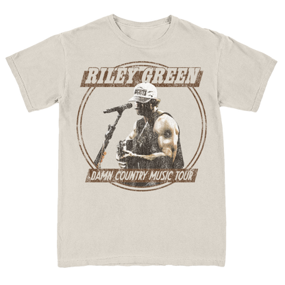 A tan-colored t-shirt with a graphic print of a person singing into a microphone and the text 'Damn Country Music Tour'. The shirt has a round neckline and short sleeves.