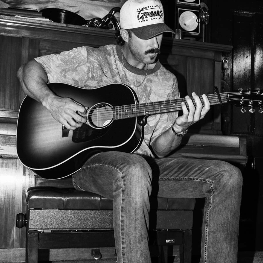 Riley Green playing an acoustic guitar in a room with wooden furniture wearing the Green's '47 hat